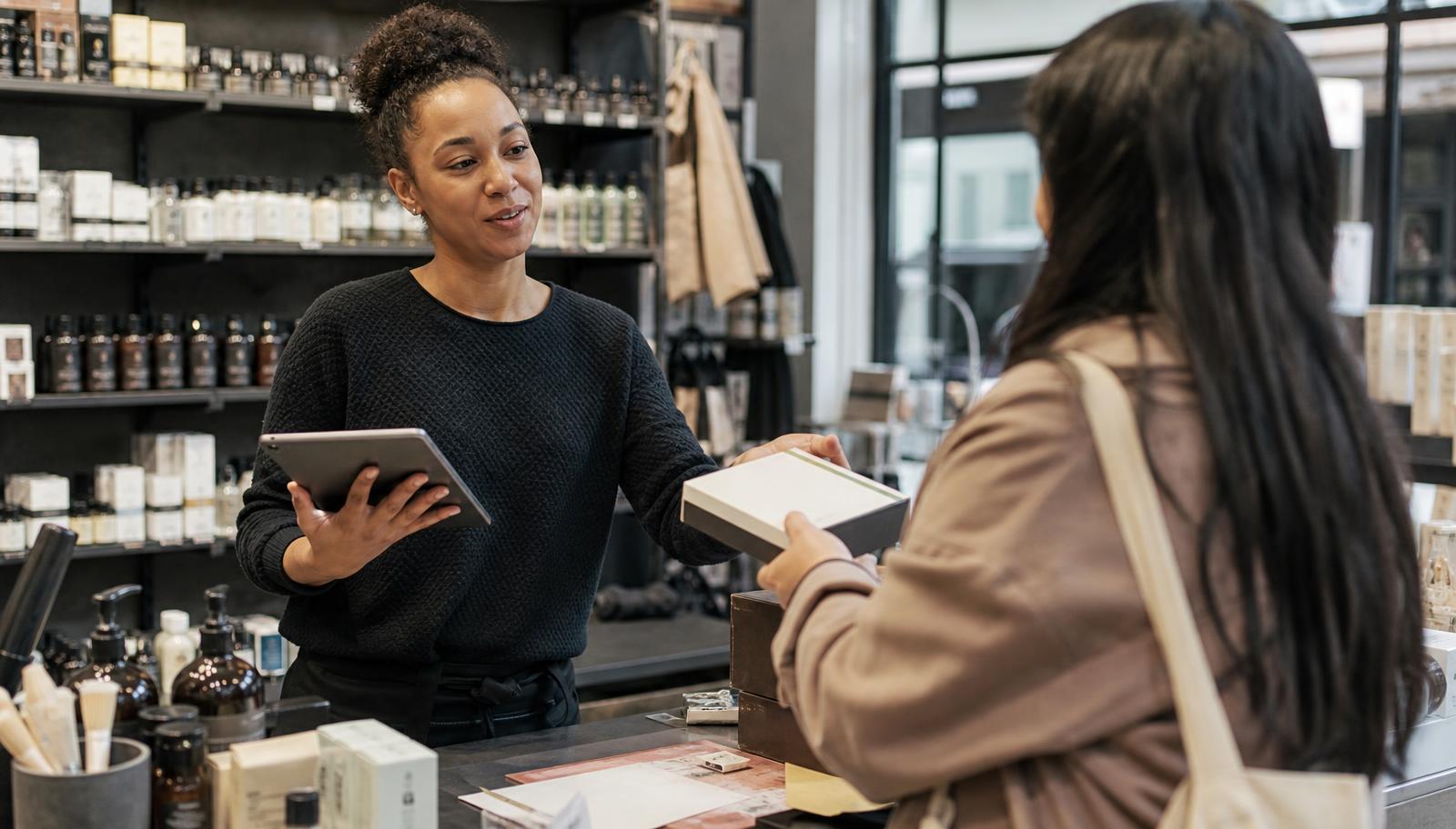 A shop assistant with a tablet handing a customer their boxed purchase — the moment a warranty is issued on Warrantik